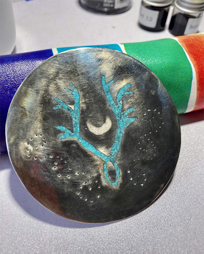 A silver-colored metal disc engraved with Nashua's clan seal, a pair of stylized antlers framing a moon, in turquoise and white inlay